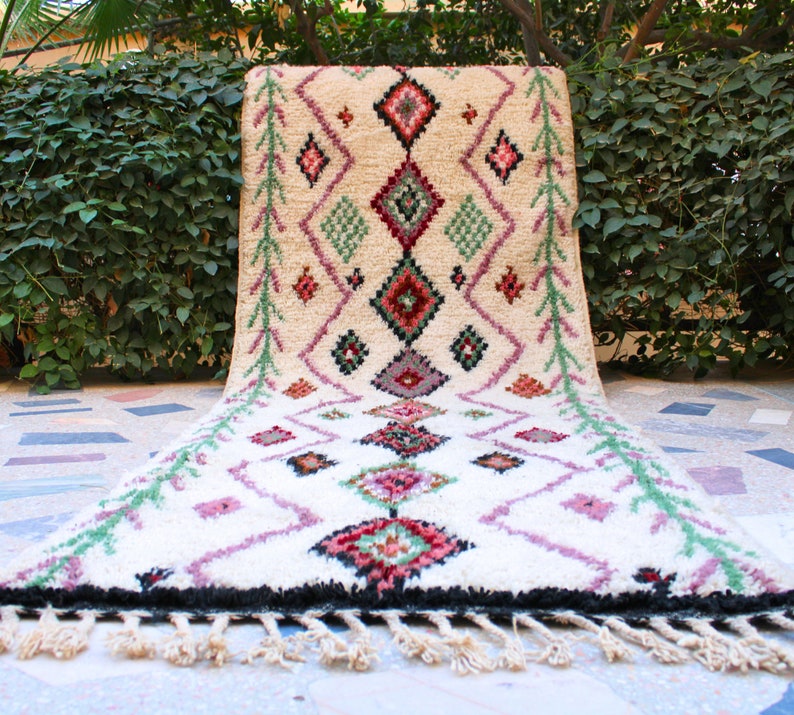 May include: A white, hand-woven rug with a geometric pattern in shades of pink, green, and red. The rug has a fringe border and is laid out on a tiled floor.