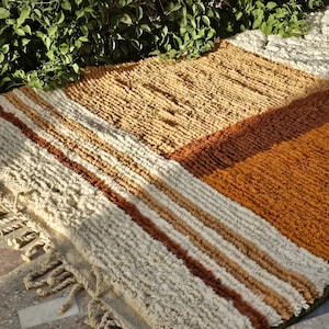 May include: A rectangular, textured rug with a geometric pattern. The rug features stripes of cream, brown, and tan, with a section of solid orange-brown. The rug has a fringe along one edge and is made of a thick, woven material.