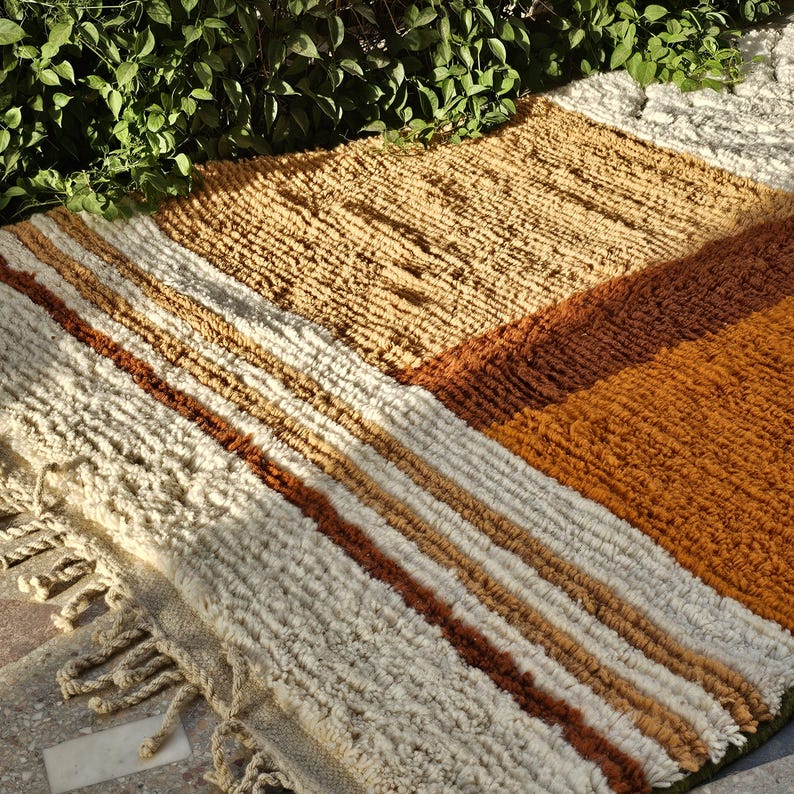 May include: A rectangular, textured rug with a geometric pattern. The rug features stripes of cream, brown, and tan, with a section of solid orange-brown. The rug has a fringe along one edge and is made of a thick, woven material.