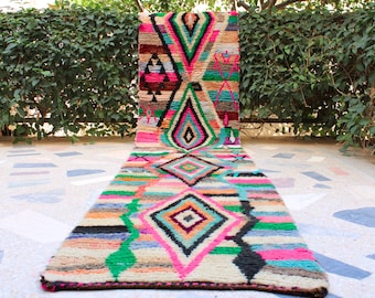 May include: A colorful Moroccan rug with a geometric pattern in shades of pink, green, blue, and black. The rug is made of wool and has a fringe on the ends.
