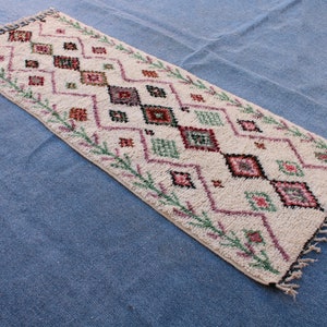 May include: A white rug with a geometric pattern in shades of pink, green, and black. The rug has a fringed border.