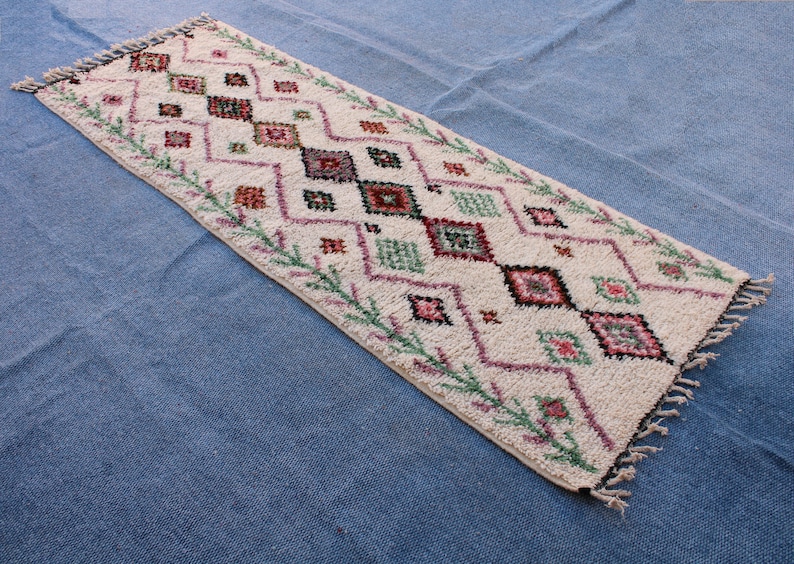 May include: A white rug with a geometric pattern in shades of pink, green, and black. The rug has a fringed border.