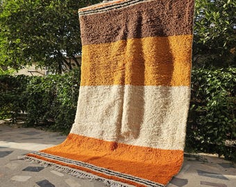 May include: A rectangular area rug featuring horizontal color-blocked stripes in shades of dark brown, mustard yellow, cream, and burnt orange. The rug has a thick, shaggy pile texture and includes thin black and white striped borders with fringe.