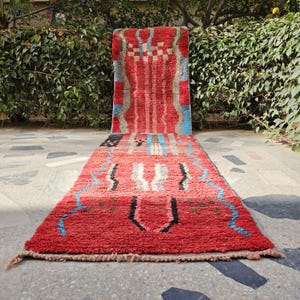 May include: A red and multi-colored runner rug with a geometric pattern. The rug features a central design with vertical stripes and a border with zig-zag and geometric shapes. The rug is made of a thick, textured material.