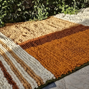May include: A rectangular rug with a textured, striped design. The rug features sections of beige, brown, and orange hues, with a green border. The rug is made of a thick, woven material.