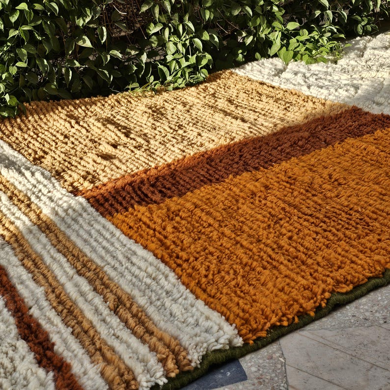 May include: A rectangular rug with a textured, striped design. The rug features sections of beige, brown, and orange hues, with a green border. The rug is made of a thick, woven material.