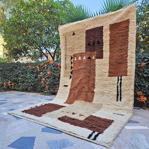 May include: A beige and brown geometric rug with a textured surface. The rug features rectangular and square shapes in varying shades of brown and black accents. The rug is displayed outdoors.