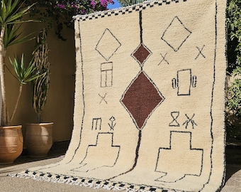 May include: Cream-colored, hand-woven rug with geometric patterns in black and brown. The rug features diamond, square, and cross-shaped designs, with a central brown diamond. The edges are trimmed with a black and white border and fringe.