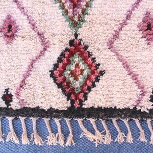 May include: A white Moroccan rug with a geometric pattern in shades of pink, green, and brown. The rug has a black border and fringe.