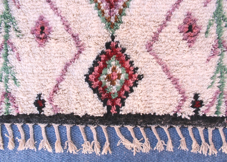 May include: A white Moroccan rug with a geometric pattern in shades of pink, green, and brown. The rug has a black border and fringe.
