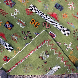 May include: A green woven rug with geometric patterns in red, white, black, and yellow. The rug has a folded corner, revealing a detailed border and additional designs. The rug's design includes various shapes and symbols.