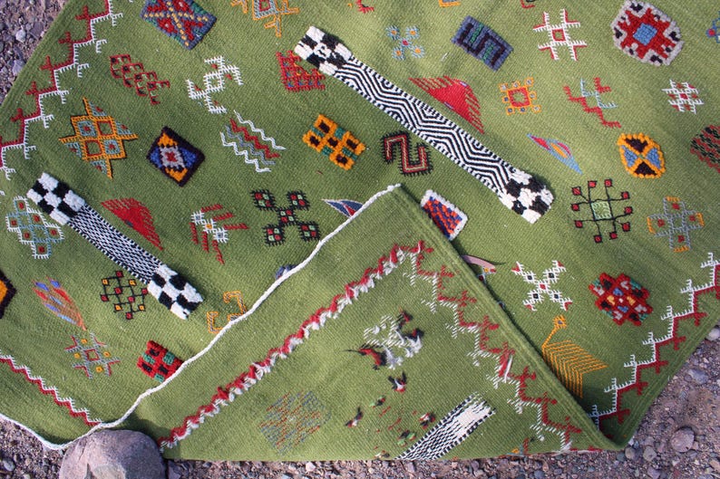 May include: A green woven rug with geometric patterns in red, white, black, and yellow. The rug has a folded corner, revealing a detailed border and additional designs. The rug's design includes various shapes and symbols.