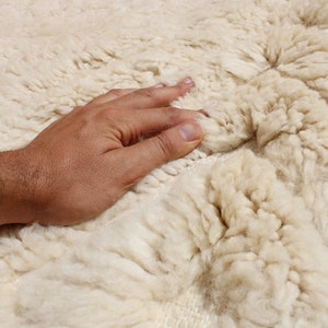 Puede incluir: Primer plano de una alfombra blanca y esponjosa con una mano tocando suavemente la superficie. La alfombra tiene un aspecto suave y texturizado.