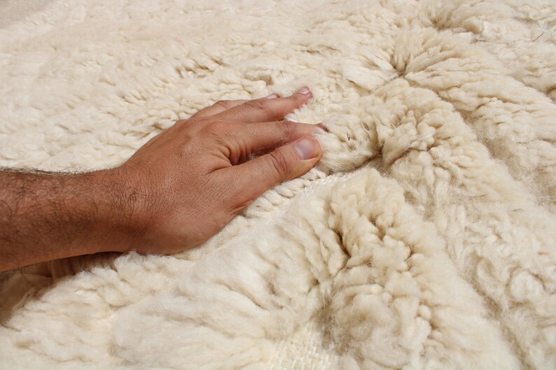Puede incluir: Primer plano de una alfombra blanca y esponjosa con una mano tocando suavemente la superficie. La alfombra tiene un aspecto suave y texturizado.