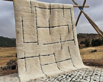 May include: A cream-colored, rectangular rug with a geometric black line design. The rug is hanging on a wooden structure outdoors, showcasing its texture and pattern. The rug's fringe is visible at the bottom.