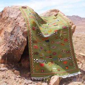 May include: A green rectangular rug with colorful geometric patterns and fringe. The rug is draped over a large rock in an outdoor setting. The rug's design includes red, white, black, and yellow accents.