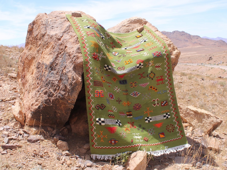 May include: A green rectangular rug with colorful geometric patterns and fringe. The rug is draped over a large rock in an outdoor setting. The rug's design includes red, white, black, and yellow accents.
