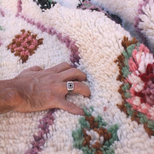 May include: A hand rests on a white, fluffy rug with a colourful Moroccan design. The rug features pink, green, and brown floral patterns. A silver ring with a black stone is on the index finger.