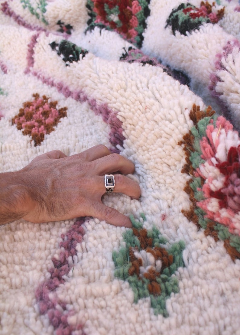 May include: A hand rests on a white, fluffy rug with a colourful Moroccan design. The rug features pink, green, and brown floral patterns. A silver ring with a black stone is on the index finger.