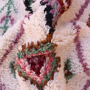 May include: Close-up of a white, fluffy rug with a colourful geometric pattern. The pattern features pink, green, brown, and black accents. The rug is made of wool and has a soft, plush texture.