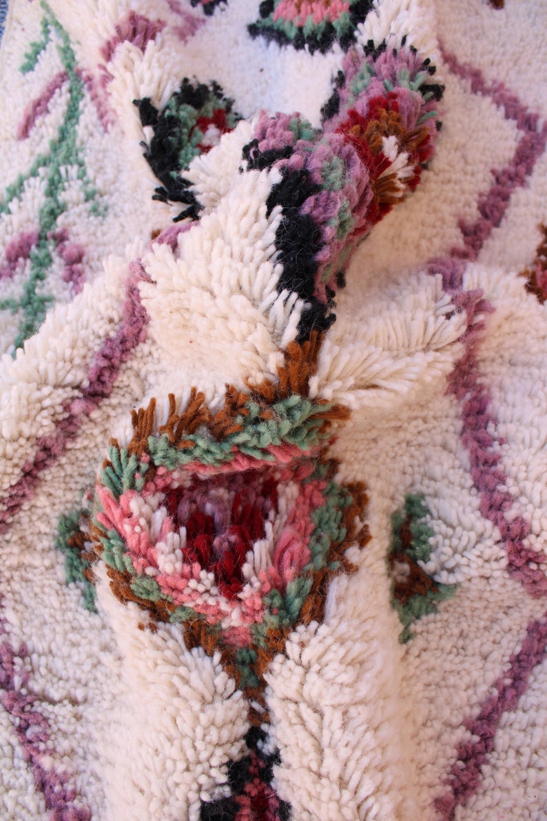 May include: Close-up of a white, fluffy rug with a colourful geometric pattern. The pattern features pink, green, brown, and black accents. The rug is made of wool and has a soft, plush texture.