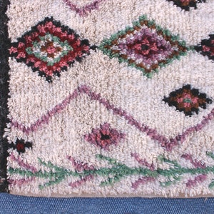 May include: A white Moroccan rug with a geometric pattern in shades of pink, green, and brown. The rug has a fringe border.