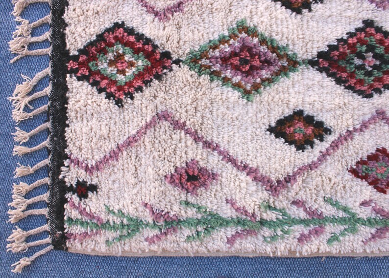 May include: A white Moroccan rug with a geometric pattern in shades of pink, green, and brown. The rug has a fringe border.