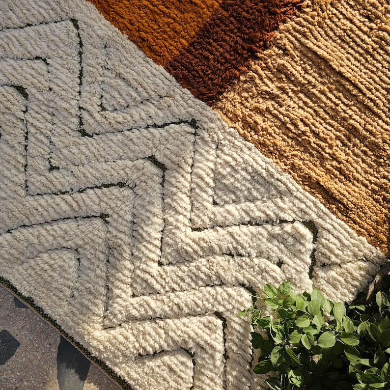 May include: Close-up of a textured rug with geometric patterns. The rug features a cream-colored zig-zag design, complemented by sections of brown and tan hues. The rug's texture is visible, with a soft, woven appearance. A small green plant is in the corner.