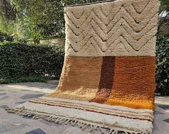 May include: A rectangular, textured rug with a geometric design. The top section features a cream-colored background with a zig-zag pattern in a darker shade. The lower section has vertical stripes in shades of brown and orange. The rug has a fringe along the bottom edge.