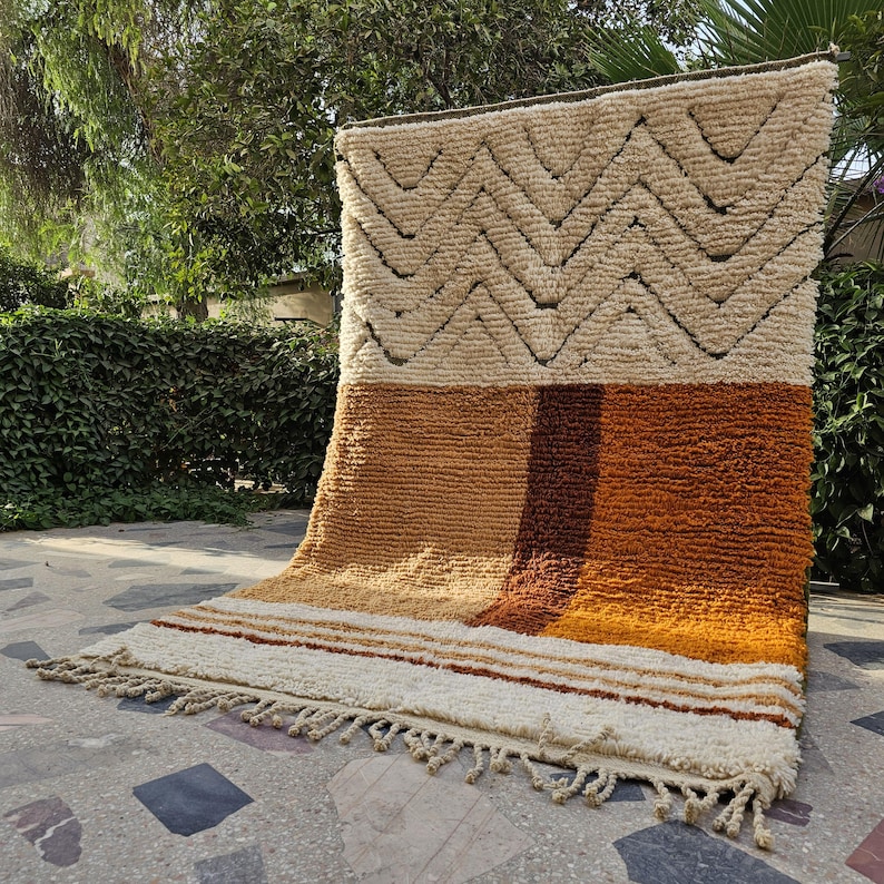 May include: A rectangular, textured rug with a geometric design. The top section features a cream-colored background with a zig-zag pattern in a darker shade. The lower section has vertical stripes in shades of brown and orange. The rug has a fringe along the bottom edge.