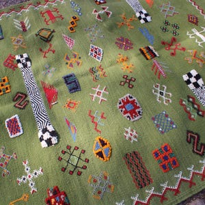 May include: A green rug with a repeating pattern of colorful geometric shapes in red, blue, yellow, and white. The rug has a fringe border and two black and white patterned bands.