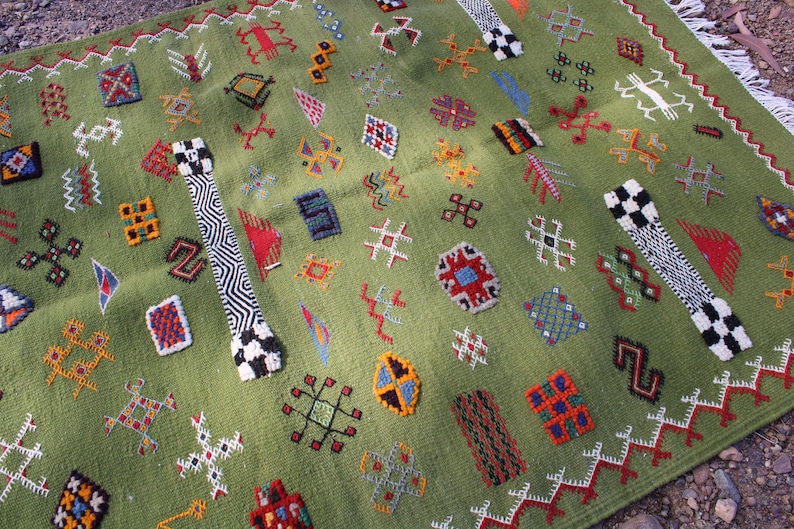 May include: A green rug with a repeating pattern of colorful geometric shapes in red, blue, yellow, and white. The rug has a fringe border and two black and white patterned bands.