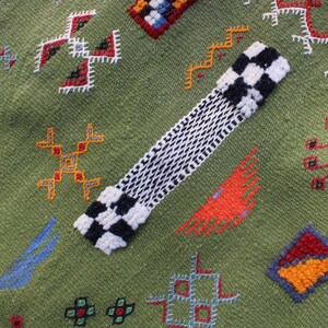 May include: A green textile with colorful embroidered geometric patterns. A black and white checkered design is on the top left. The textile has red, orange, blue, and yellow accents.