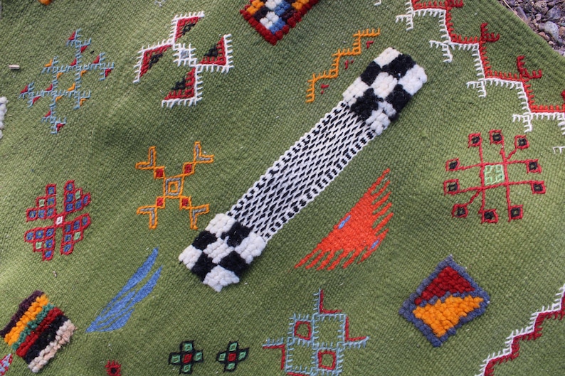 May include: A green textile with colorful embroidered geometric patterns. A black and white checkered design is on the top left. The textile has red, orange, blue, and yellow accents.