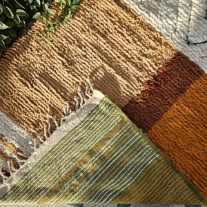 May include: A textured, multi-colored rug with a geometric design. The rug features sections of tan, brown, and orange, with a green and white striped underside. The edges have a fringe detail. This rug is suitable for home decor.