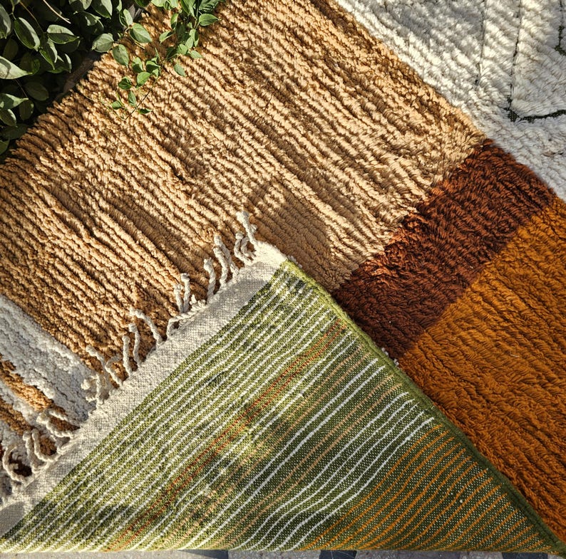 May include: A textured, multi-colored rug with a geometric design. The rug features sections of tan, brown, and orange, with a green and white striped underside. The edges have a fringe detail. This rug is suitable for home decor.