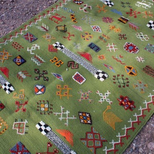 May include: A green rectangular rug with a detailed pattern of various shapes and symbols in red, orange, blue, and white. The rug has a fringe on one end and is placed on a textured surface.