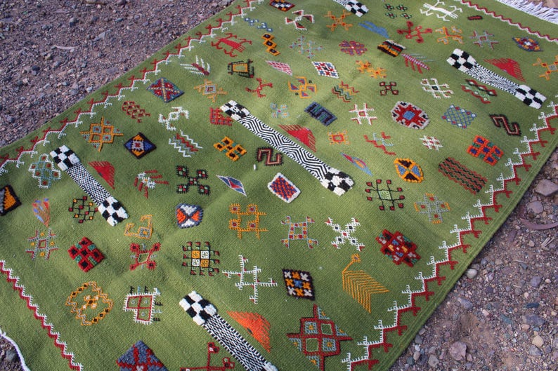 May include: A green rectangular rug with a detailed pattern of various shapes and symbols in red, orange, blue, and white. The rug has a fringe on one end and is placed on a textured surface.