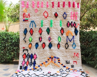 May include: A white Moroccan rug with a geometric pattern in bright colors, including pink, blue, green, orange, and red. The rug has a white fringe border.