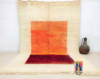 May include: A white Moroccan rug with a large orange and red square in the center. The rug has a fringe border.