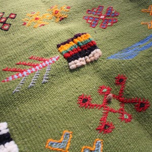 May include: Close-up of a green textile with colorful embroidered patterns. The design includes geometric shapes and floral motifs in red, yellow, blue, and black. The texture of the fabric is visible, showcasing the detailed craftsmanship.