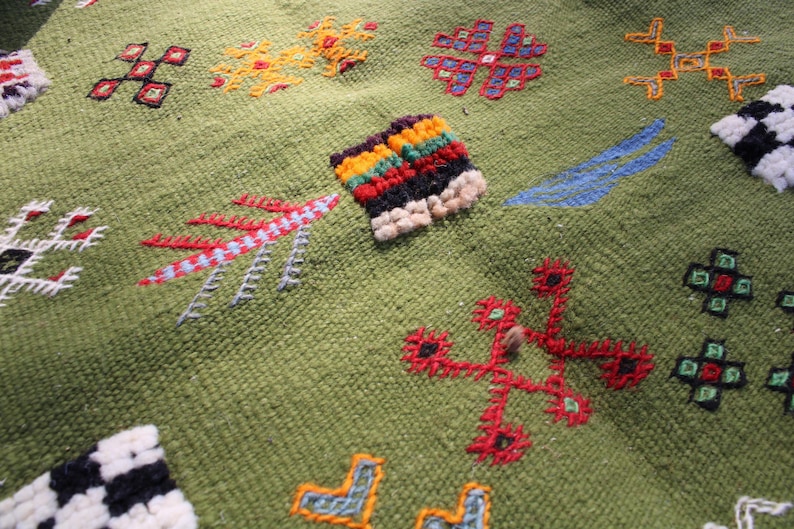 May include: Close-up of a green textile with colorful embroidered patterns. The design includes geometric shapes and floral motifs in red, yellow, blue, and black. The texture of the fabric is visible, showcasing the detailed craftsmanship.