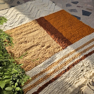May include: A textured, rectangular rug with a geometric design. The rug features sections of cream, tan, brown, and rust colors. The rug has a shaggy, woven texture and is suitable for a living room or bedroom.