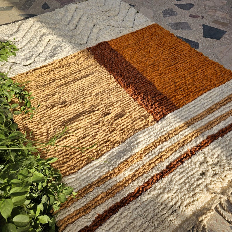 May include: A textured, rectangular rug with a geometric design. The rug features sections of cream, tan, brown, and rust colors. The rug has a shaggy, woven texture and is suitable for a living room or bedroom.