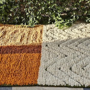 May include: A rectangular rug with a textured, geometric design. The rug features sections of varying colors, including shades of gold, brown, and cream. The rug is made of a thick, woven material.