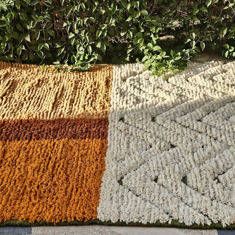 May include: A rectangular rug with a textured, geometric design. The rug features sections of varying colors, including shades of gold, brown, and cream. The rug is made of a thick, woven material.