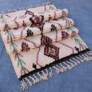 May include: A stack of three white Moroccan rugs with pink, green and black geometric patterns. The rugs have fringe on the bottom.