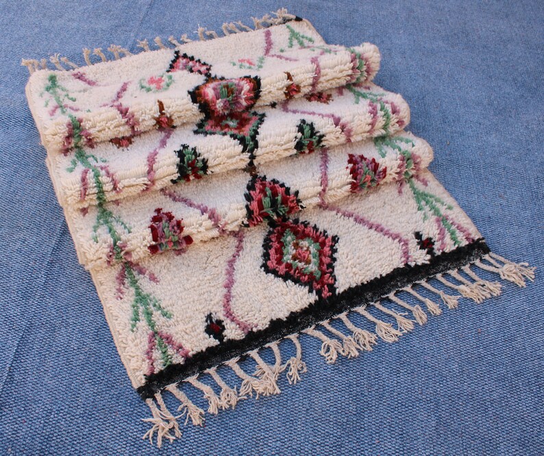 May include: A stack of three white Moroccan rugs with pink, green and black geometric patterns. The rugs have fringe on the bottom.