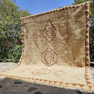 May include: A large, rectangular, cream-colored shag rug featuring a thick, high-pile texture. The rug is decorated with brown geometric diamond and square motifs arranged in a symmetrical pattern, finished with a brown triangular border.