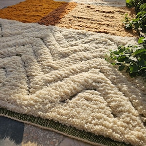 May include: A textured, geometric rug with a cream, tan, and orange color scheme. The rug features a repeating pattern of cream-colored shapes against a tan background, with a border of green. The rug is made of a soft, plush material.
